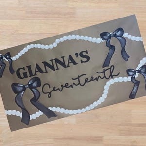 Op de afbeelding: Een bruin rechthoekig bord met de tekst "GIANNA'S Seventeenth" in zwart schrift. Het bord is versierd met zwarte strikken en witte parelachtige accenten. De strikken zijn in een donkergrijze kleur geschilderd.