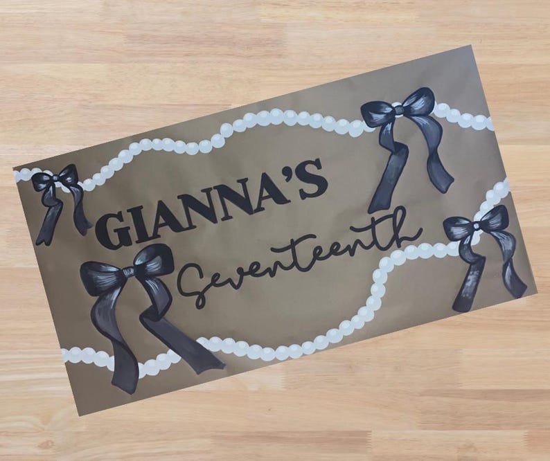 Op de afbeelding: Een bruin rechthoekig bord met de tekst "GIANNA'S Seventeenth" in zwart schrift. Het bord is versierd met zwarte strikken en witte parelachtige accenten. De strikken zijn in een donkergrijze kleur geschilderd.