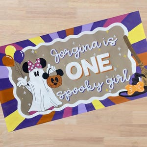 Op de afbeelding: Een rechthoekig, handgeschilderd bord met een Halloween-thema. Het bord toont een geest verkleed als Minnie Mouse met een pompoen en ballonnen. De tekst luidt "Jorgina is ONE spooky girl" in witte letters. Een heksenhoed en een bezem zijn ook afgebeeld.