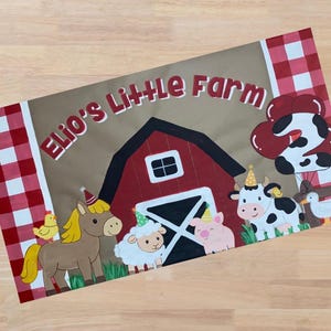 Op de afbeelding: Een rechthoekig bord met de tekst "Elio's Little Farm" boven een rode schuurillustratie. Tekenfilm dieren, waaronder een paard, een schaap, een varken, een koe en een eend, dragen feesthoeden. Rood-witte geblokte randen omlijsten het ontwerp.