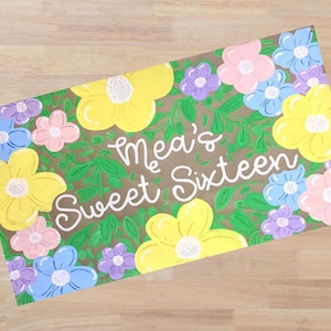 Op de afbeelding: Een rechthoekig bord met de tekst "Mea's Sweet Sixteen" in witte letters, omringd door kleurrijke bloemen. De bloemen zijn geel, roze, blauw en paars, tegen een groene, bladerrijke achtergrond.