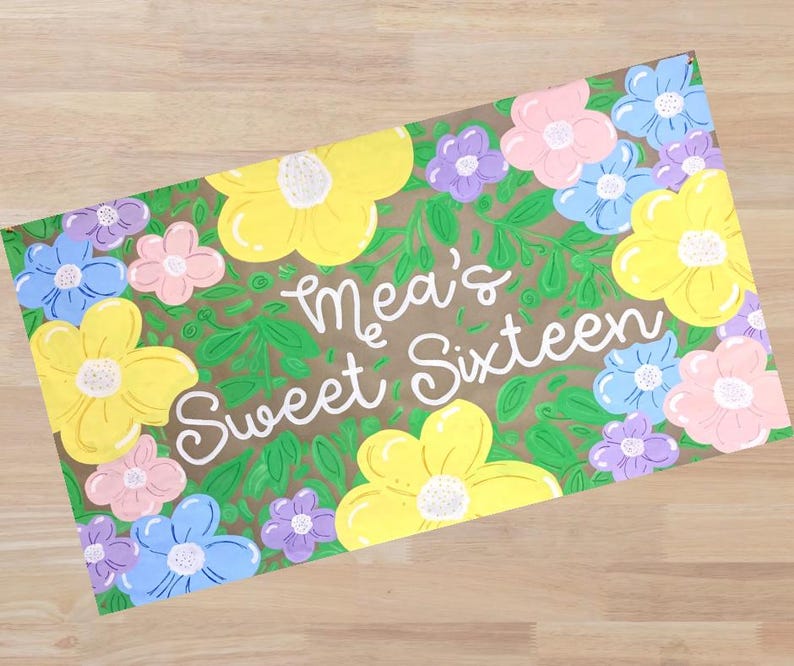 Op de afbeelding: Een rechthoekig bord met de tekst "Mea's Sweet Sixteen" in witte letters, omringd door kleurrijke bloemen. De bloemen zijn geel, roze, blauw en paars, tegen een groene, bladerrijke achtergrond.