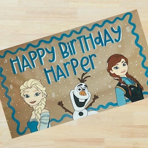 Op de afbeelding: Handgeschilderde verjaardagskaart op bruin papier met de woorden "Happy Birthday Harper" in blauw. De kaart bevat illustraties van Elsa, Anna en Olaf uit de film Frozen, met een blauwe golvende rand.