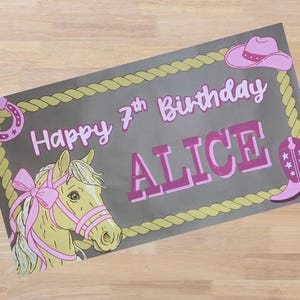 Op de afbeelding: Rechthoekige banner met een grijze achtergrond, met een paardenkop, hoefijzer, cowboyhoed en laars. De banner leest "Happy 7th Birthday ALICE" in roze letters, omlijst door een touwrand.
