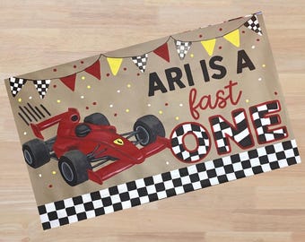 May include: A rectangular sign on a tan background featuring a red race car, a black and white checkered border, and colorful pennant flags. The text reads ARI IS A fast ONE in black and red lettering with a checkered pattern on the word ONE.