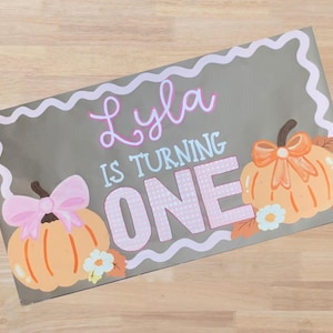 Op de afbeelding: Een grijs rechthoekig bord met de tekst "Lyla IS TURNING ONE" in roze en wit. Het bord heeft twee oranje pompoenen met strikken en bloemaccenten, en een witte golvende rand.