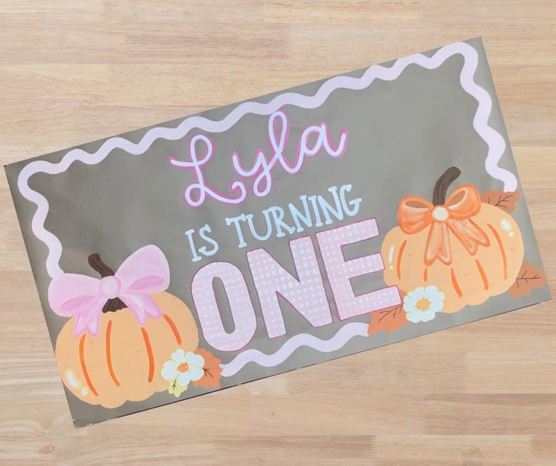 Op de afbeelding: Een grijs rechthoekig bord met de tekst "Lyla IS TURNING ONE" in roze en wit. Het bord heeft twee oranje pompoenen met strikken en bloemaccenten, en een witte golvende rand.
