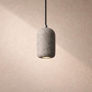 May include: A gray, cylindrical pendant light fixture with a textured surface hangs from a black cord. The light fixture is illuminated from within, casting a warm glow. The background is a light beige color.