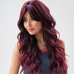 May include: A woman with long, wavy, burgundy-colored hair. The hair has a side part and bangs. She is wearing a navy blue tank top with a red and white trim and a gold necklace.