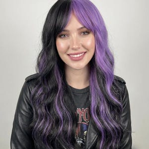 May include: A long, wavy wig with a split design, featuring black hair on one side and vibrant purple on the other. The wig has a side bang and is worn by a person wearing a black leather jacket. The text "Vision Wigs" is visible.