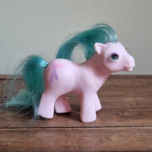 Puede incluir: Un poni de juguete rosa vintage con pelo y cola turquesa. El poni tiene un diseño floral morado en un costado y está sobre una superficie de madera. Los ojos del poni son de color azul claro.