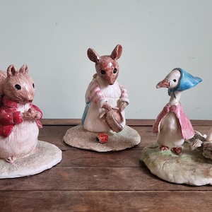 Puede incluir: Tres figuras de cerámica de personajes de cuentos. Una ardilla con una chaqueta roja, un ratón con un vestido y un ganso con un gorro azul y una capa rosa. Cada figura está sobre una pequeña base texturizada, colocada sobre una superficie de madera.