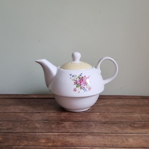 May include: White ceramic teapot with a floral design. The teapot has a rounded body, a curved spout, and a loop handle. The lid is yellow with a small knob. The teapot is decorated with a cluster of pink and purple flowers.