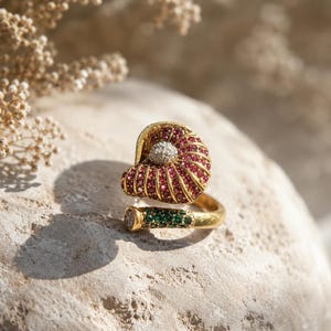 May include: A gold ring featuring a shell-shaped top encrusted with small red stones and a central white stone cluster. The band is open-ended, with one side decorated with green stones and a small white stone accent at the very tip of the metal.