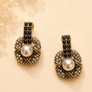 May include: A pair of drop earrings featuring a central white faux pearl surrounded by a square frame of clear crystals and antique gold-toned metal. The top section of each earring is a vertical bar encrusted with small black crystals.