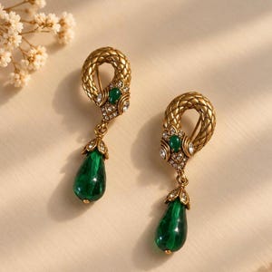 May include: A pair of drop earrings featuring a textured gold-tone snake motif coiled into a loop. Each earring is accented with a small green stone and clear crystals, finished with a dangling, pear-shaped deep green glass teardrop pendant.