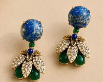 May include: A pair of drop earrings featuring a large round blue lapis lazuli stone at the top, connected to a cluster of gold-tone metal leaves encrusted with clear crystals and two smooth, teardrop-shaped green stones at the base.