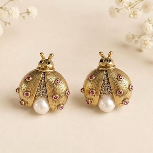 May include: A pair of gold-toned ladybird earrings featuring a textured metal finish, small pink circular crystals embedded on the wings, a central cluster of clear rhinestones, and a single white pearl accent at the base of each insect body.