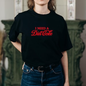 May include: Black t-shirt with the red text "I NEED A Diet Coke" in a stylized font. The shirt is paired with blue jeans and a black belt. The overall style is casual and comfortable.