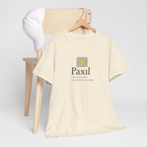 Può includere: T-shirt color crema con la parola "Paxil" in un font stilizzato, accompagnata da testo sottostante. Una piccola grafica quadrata con un disegno di sole è sopra la parola. Un berretto da baseball bianco è appoggiato su una sedia di legno.