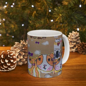 May include: White ceramic mug featuring a colorful, hand-drawn design of cartoon dogs with orange and white fur. The mug has a brown background with drawings of clouds, butterflies, and the name "Maria Lima". The mug is on a wooden surface.