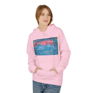 Pode incluir: Um moletom rosa claro com um bolso frontal. O moletom apresenta uma pintura infantil e colorida de uma paisagem com uma estrutura vermelha, água azul e árvores verdes. A modelo está usando jeans azuis.