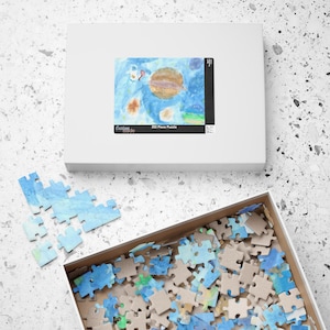 Pode incluir: Uma caixa de quebra-cabeça branca com uma imagem colorida com tema espacial. A caixa está rotulada como "252 Piece Puzzle". Peças de quebra-cabeça espalhadas são visíveis, juntamente com uma caixa aberta cheia de mais peças. A obra de arte apresenta um planeta e outros elementos celestes.