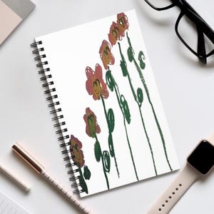 May include: A white spiral-bound notebook with a floral illustration in red, green, and yellow. The notebook is open, revealing the artwork. A pen, glasses, and a smartwatch are in the background, suggesting a workspace or desk.