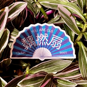 May include: A fan-shaped sticker with a blue and purple design, featuring the text "YOU ARE OLS" and Chinese characters. The sticker also includes the words "chi lun" and "seen". The sticker is set against a backdrop of green and purple-striped leaves.