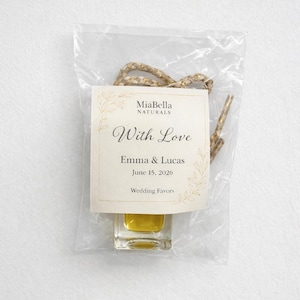 Op de afbeelding: Een kleine, heldere glazen fles gevuld met een gele vloeistof, verpakt in een doorzichtige plastic zak. Een crèmekleurig etiket leest "MiaBella NATURALS" en "With Love" met de namen Emma & Lucas en de datum 15 juni 2026. Bruiloft bedankjes.