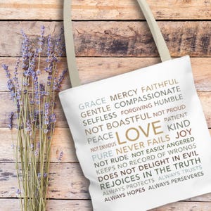 May include: A white tote bag featuring the word "LOVE" in gold and inspirational text. The bag has beige straps and is next to a bundle of lavender. The text includes words like "grace", "mercy", and "joy".