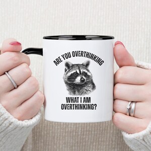 May include: White ceramic mug with a black handle and rim. The mug features a black and white illustration of a raccoon and the text "ARE YOU OVERTHINKING WHAT I AM OVERTHINKING?"