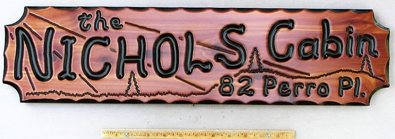 May include: A wooden sign with a carved design of a mountain range and trees. The sign reads "the NICHOLS Cabin 82 Perro Pl."
