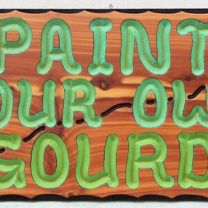 May include: A wooden sign with the words "PAINT YOUR OWN GOURD" in green raised letters. The sign is decorated with a carved gourd and vine design.