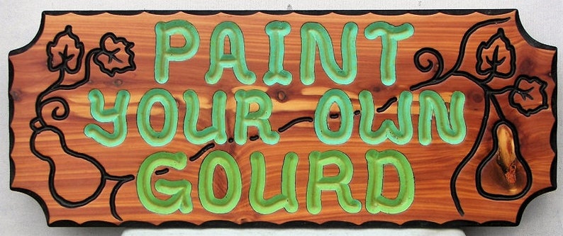 May include: A wooden sign with the words "PAINT YOUR OWN GOURD" in green raised letters. The sign is decorated with a carved gourd and vine design.