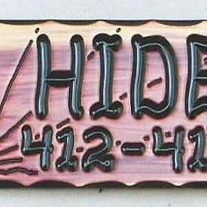 May include: A wooden sign with a rustic, distressed look. The sign reads "THE HIDEOUT" in black lettering with a stylized font. Below the text is a sunrise graphic and the numbers "412-418-9478".
