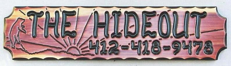 May include: A wooden sign with a rustic, distressed look. The sign reads "THE HIDEOUT" in black lettering with a stylized font. Below the text is a sunrise graphic and the numbers "412-418-9478".