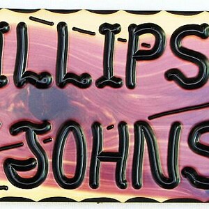 May include: A wooden sign with a red-brown stain and black lettering. The sign reads "Phillips & Johnson" with a palm tree graphic on the left and a sunburst graphic on the right.