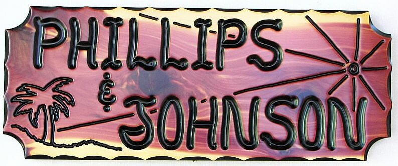 May include: A wooden sign with a red-brown stain and black lettering. The sign reads "Phillips & Johnson" with a palm tree graphic on the left and a sunburst graphic on the right.