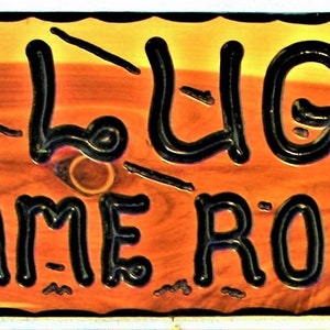 May include: A wooden sign with a black border and black lettering that reads "A.G.L.UGO'S GAME ROOM". The sign has a rustic, wood-grain texture.
