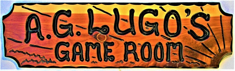 May include: A wooden sign with a black border and black lettering that reads "A.G.L.UGO'S GAME ROOM". The sign has a rustic, wood-grain texture.