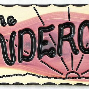 May include: A wooden sign with a rustic, reddish-brown background and a black border. The sign reads "the Ponderosa" in a stylized, black font. The sign also features a stylized sunrise and a small tree on the right side.