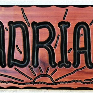 May include: A wooden sign with a rustic, reddish-brown finish and a black border. The sign has a wavy, decorative edge. The text "MADRI&Aacute;N'S" is carved in black lettering with a stylized, whimsical font. The sign features a stylized sunrise graphic below the text.