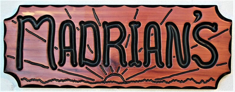 May include: A wooden sign with a rustic, reddish-brown finish and a black border. The sign has a wavy, decorative edge. The text "MADRI&Aacute;N'S" is carved in black lettering with a stylized, whimsical font. The sign features a stylized sunrise graphic below the text.
