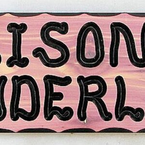 May include: A pink wooden sign with black lettering that reads "Alison's Wonderland". The sign has a scalloped edge and two carved oak leaves on either side of the text.