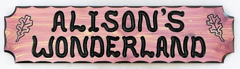 May include: A pink wooden sign with black lettering that reads "Alison's Wonderland". The sign has a scalloped edge and two carved oak leaves on either side of the text.