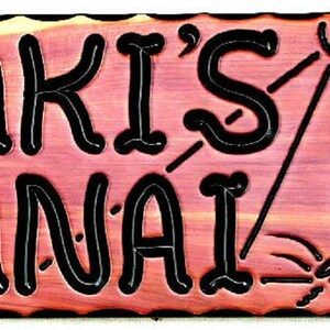 May include: A wooden sign with the text "Laki's Lanai" in black lettering. The sign has a carved image of a fish jumping out of the water with the sun shining above.