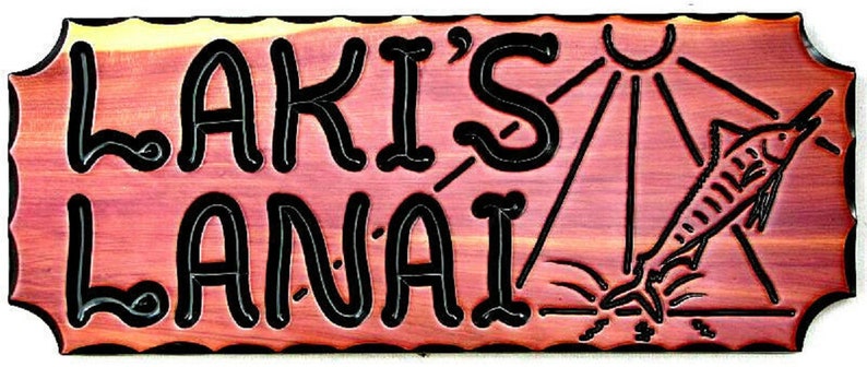 May include: A wooden sign with the text "Laki's Lanai" in black lettering. The sign has a carved image of a fish jumping out of the water with the sun shining above.