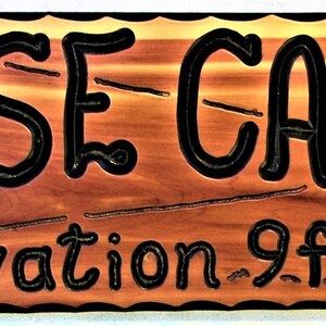 May include: A wooden sign with the words "BASE CAMP" in black lettering on a reddish-brown background. The sign also includes the text "Elevation 9 ft" in black lettering.