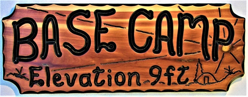 May include: A wooden sign with the words "BASE CAMP" in black lettering on a reddish-brown background. The sign also includes the text "Elevation 9 ft" in black lettering.